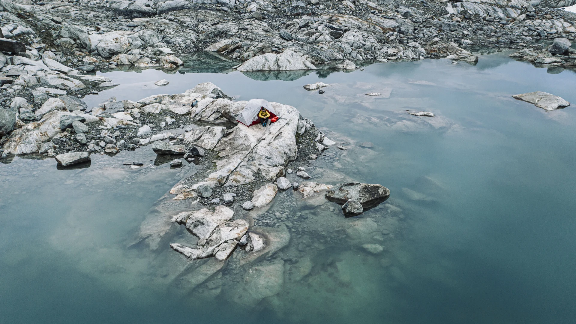 Small tent on rocks beside a still mountain lake