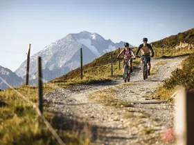 Two cyclists on a mountain trail with snow-capped peaks in the background